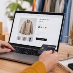 Profesional realizando una compra online en un entorno de retail y ecommerce con portátil, carrito de compra, paquetes y elementos de marketing digital
